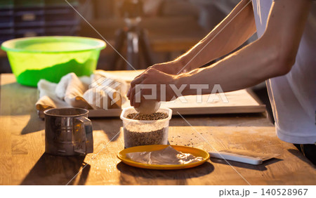 Skilled baker hands meticulously prepare dough, rolling ball in mixture of seeds on rustic wooden table. Golden light highlights culinary craftsmanship during baking process. 140528967