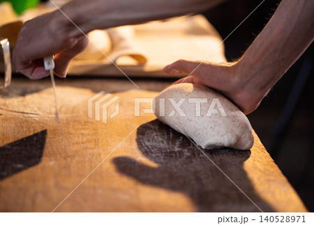 Skilled hands of baker carefully shaping fresh dough loaf on wooden surface dusted with flour preparation for baking bread Skilled hands of baker carefully shaping fresh dough loaf on wooden surface dusted with flour preparation for baking bread 140528971