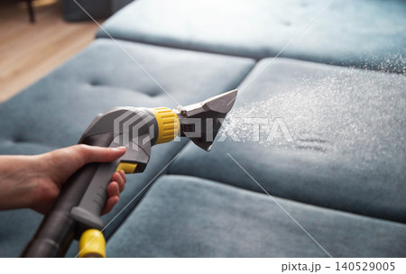 Close-up of hand holding upholstery cleaning tool spraying fine mist onto blue fabric sofa during deep cleaning process in modern home environment Close-up of hand holding upholstery cleaning tool spraying fine mist onto blue fabric sofa during deep cleaning process in modern home environment 140529005
