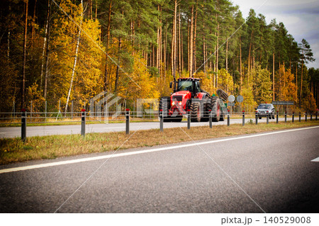 Red agricultural tractor with attached trailer driving on asphalt road, green roadside barrier, vibrant autumn trees and evergreen forest lining background, black SUV passing opposite lane, rural 140529008
