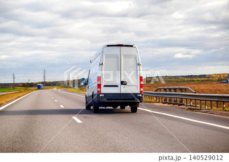 White cargo van driving on highway under cloudy sky during day, transport vehicle moving on asphalt road with fields and trees in background White cargo van driving on highway under cloudy sky during day, transport vehicle moving on asphalt road with fields and trees in background 140529012