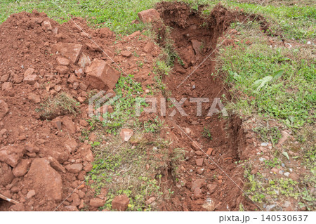 A high angle view of an open earthworks trench split junction dug into dark brown soil and green lawn grass for multi-line residential utility layout. Underground infrastructure pipe intersection 140530637