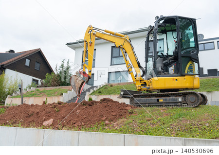 Tracked yellow mini excavator working on a grassy hillside plot above a new concrete retaining wall foundation during home renovation. Residential landscaping project showing a compact digger leveling 140530696