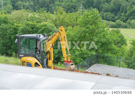 A yellow mini excavator operating on a countryside domestic property during garden remodeling and terrain earthworks. Compact industrial construction digger vehicle standing on a hill slope site A yellow mini excavator operating on a countryside domestic property during garden remodeling and terrain earthworks. Compact industrial construction digger vehicle standing on a hill slope site 140530698