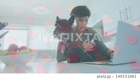 Holding terrier, woman in longsleeve top typing on laptop at kitchen counter, pink heart overlay Holding terrier, woman in longsleeve top typing on laptop at kitchen counter, pink heart overlay 140531061