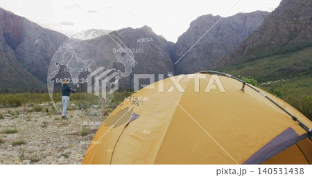 Walking camper in dark jacket holding phone toward pass at mountain valley, yellow tent, AR overlay Walking camper in dark jacket holding phone toward pass at mountain valley, yellow tent, AR overlay 140531438