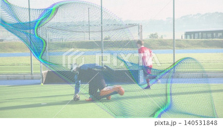 Kneeling goalkeeper in helmet and glove reaching for ball on synthetic turf pink-clad player racing 140531848