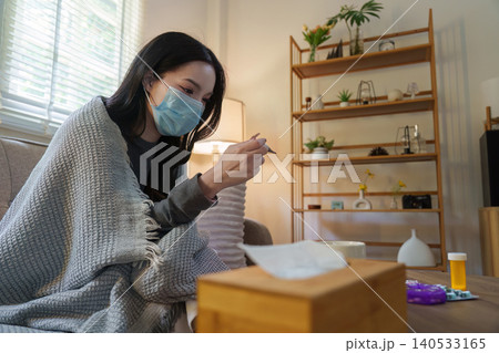 Asian woman with face mask checking temperature with thermometer at home Asian woman with face mask checking temperature with thermometer at home 140533165
