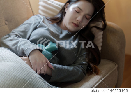 Asian woman resting on couch with hot water bottle for period pain relief 140533166