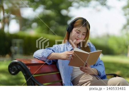 Asian woman reading book with headphones on park bench, self-care 140533481