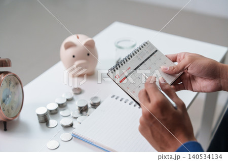 Asian man planning schedule with calendar, pen, and savings on desk Asian man planning schedule with calendar, pen, and savings on desk 140534134