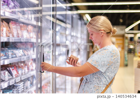 Woman choosing frozen food from supermarket refrigerator aisle while reading product label and opening glass freezer door in modern grocery store Woman choosing frozen food from supermarket refrigerator aisle while reading product label and opening glass freezer door in modern grocery store 140534634