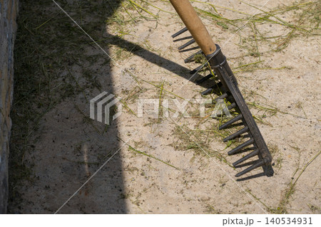 A metal garden rake with a wooden handle rests on dry sandy ground scattered with grass clippings, casting a strong shadow in bright sunlight 140534931