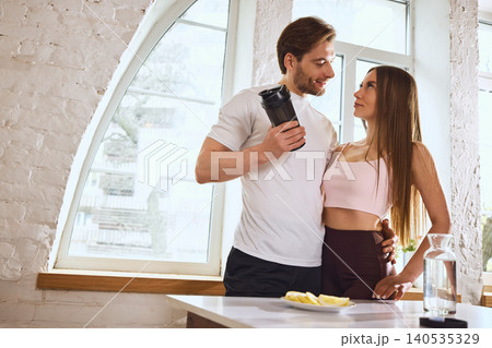 Fit couple standing together with protein shaker and lemon water in bright home kitchen 140535329