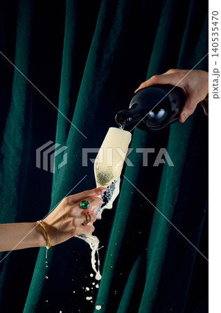 Champagne overflowing from glass held by hand with foam splash on dark green velvet background Champagne overflowing from glass held by hand with foam splash on dark green velvet background 140535470
