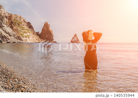 Woman sea coast standing in clear water near rocks wearing a black dress under a warm sunset glow 140535944