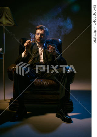 Confident man in suit smoking cigar and holding whiskey in dramatic low key lighting. Confident man in suit smoking cigar and holding whiskey in dramatic low key lighting. 140536048