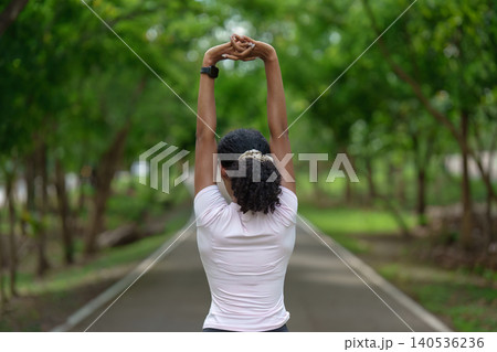 Rear view of Black woman stretching arms overhead on park path for outdoor fitness Rear view of Black woman stretching arms overhead on park path for outdoor fitness 140536236