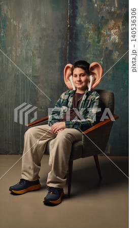 Boy with oversized ears and earbuds sits calmly in chair against textured wall. 140536306