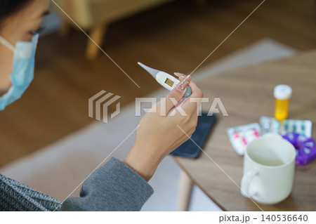 Asian woman in medical mask checking temperature with thermometer at home for illness Asian woman in medical mask checking temperature with thermometer at home for illness 140536640