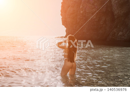 Woman sunset ocean waves reflect golden light as a female in bikini stands in a serene coastal cove during summer 140536976
