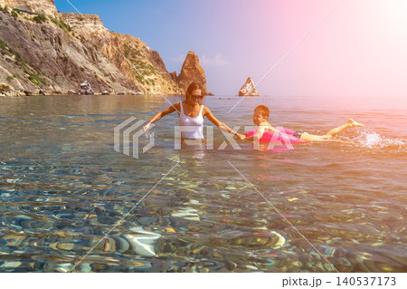 Family beach vacation mother and son swimming in crystal clear sea water near rocky cliffs on sunny day Family beach vacation mother and son swimming in crystal clear sea water near rocky cliffs on sunny day 140537173