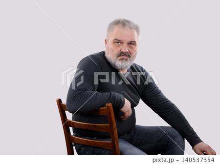 Serious mature bearded man sitting on chair and looking at camera Serious mature bearded man sitting on chair and looking at camera 140537354