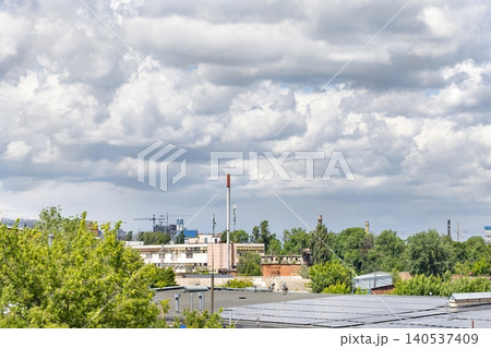 Cloudy sky over industrial area with buildings, trees, and a smokestack in the background during daytime 140537409