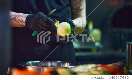 Cook peeling onion with knife over metal bowl in professional kitchen. 140537486