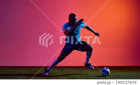 Soccer player sprinting with ball in blue uniform on pink studio field Soccer player sprinting with ball in blue uniform on pink studio field 140537639