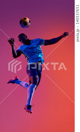 Soccer player heading ball in blue sport uniform on pink studio field. Soccer player heading ball in blue sport uniform on pink studio field. 140537651