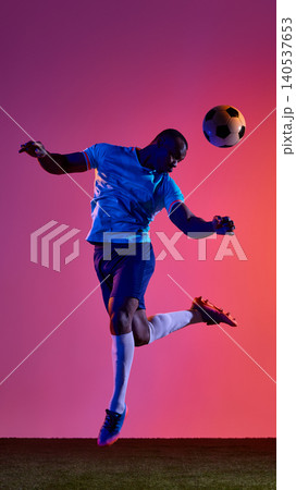 Football player jumping to head ball on grass field against pink orange background. Football player jumping to head ball on grass field against pink orange background. 140537653