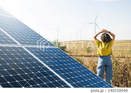 Rear view of a woman observing wind turbines. Solar panel in the foreground 140538040