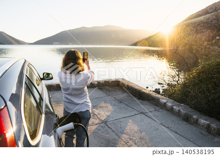 Woman with smartphone next to charging electric car on the seacost during summer vacation. 140538195