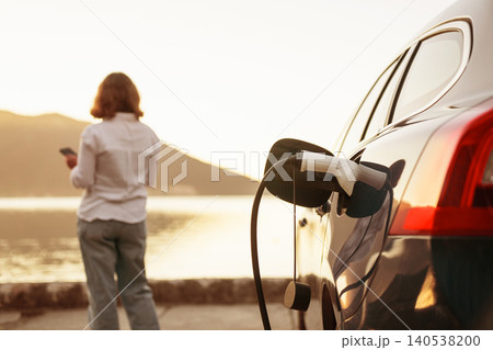 Woman with smartphone next to charging electric car on the seacost during summer vacation. Woman with smartphone next to charging electric car on the seacost during summer vacation. 140538200