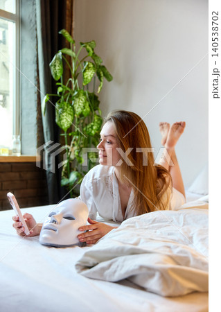 Woman browses smartphone in bed beside LED facial mask in cozy home setting with natural light Woman browses smartphone in bed beside LED facial mask in cozy home setting with natural light 140538702
