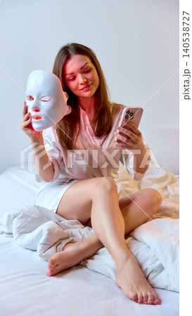 Woman checks smartphone while holding LED facial mask in bed, with soft natural light, white bedding, and clean home setting. Woman checks smartphone while holding LED facial mask in bed, with soft natural light, white bedding, and clean home setting. 140538727
