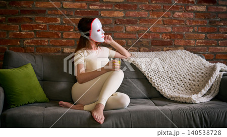 Woman wears LED facial mask on sofa and holds glass of water, with warm home light, brick wall background, and relaxed composition Woman wears LED facial mask on sofa and holds glass of water, with warm home light, brick wall background, and relaxed composition 140538728