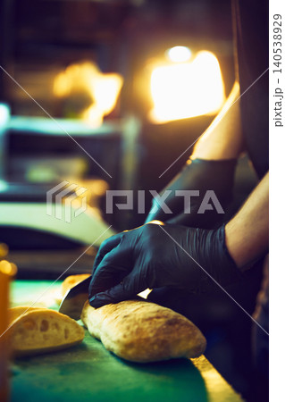 Chef cutting crusty bread under warm lights in commercial kitchen. Chef cutting crusty bread under warm lights in commercial kitchen. 140538929