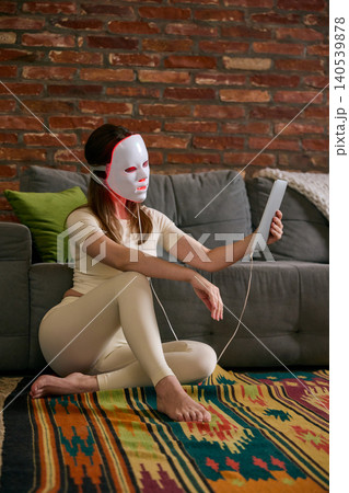 Woman sits on floor using LED face mask device near sofa, with warm indoor light, colorful rug, and candid home setting. Woman sits on floor using LED face mask device near sofa, with warm indoor light, colorful rug, and candid home setting. 140539878
