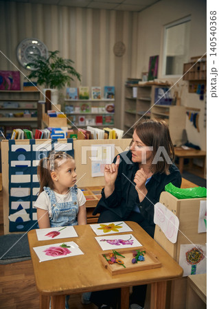 Serious girl listening to educator during flower vocabulary lesson in daycare room Serious girl listening to educator during flower vocabulary lesson in daycare room 140540368