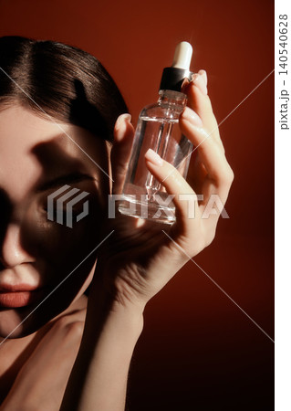 Woman holding glass serum bottle beside half shadowed face on orange background Woman holding glass serum bottle beside half shadowed face on orange background 140540628