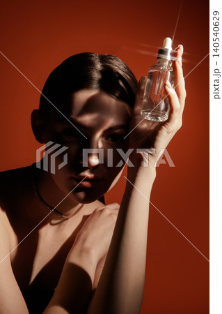 Woman holding glass serum bottle beside face under hard shadow on orange background Woman holding glass serum bottle beside face under hard shadow on orange background 140540629