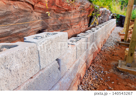 Workers place concrete blocks in row on building site surrounded by rocks during installing retaining wall Workers place concrete blocks in row on building site surrounded by rocks during installing retaining wall 140543267