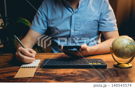 Person Using Smartphone While Writing Notes at a Wooden Desk with Globe in a Cozy Home Office Setting Person Using Smartphone While Writing Notes at a Wooden Desk with Globe in a Cozy Home Office Setting 140543574