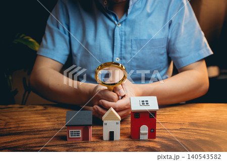 Person Examining Miniature Houses with Magnifying Glass on Wooden Table for Real Estate and Investment Concepts Person Examining Miniature Houses with Magnifying Glass on Wooden Table for Real Estate and Investment Concepts 140543582