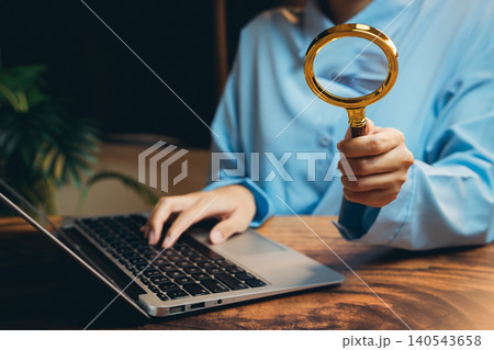 Person Using Magnifying Glass While Working on Laptop Computer in Home Office Setting with Green Plant Background Person Using Magnifying Glass While Working on Laptop Computer in Home Office Setting with Green Plant Background 140543658