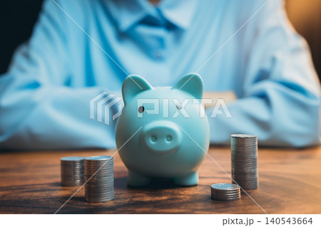 Cute blue piggy bank surrounded by stacks of coins on a wooden table with a person in the background Cute blue piggy bank surrounded by stacks of coins on a wooden table with a person in the background 140543664