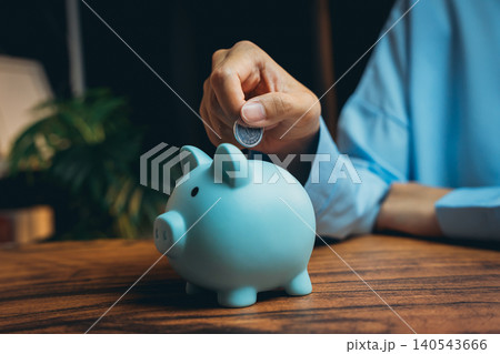 Hand Inserting Coin Into Cute Blue Piggy Bank on Wooden Table with Background of Indoor Plant for Saving Concept Hand Inserting Coin Into Cute Blue Piggy Bank on Wooden Table with Background of Indoor Plant for Saving Concept 140543666