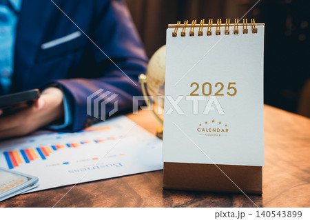 Desk Calendar for Year 2025 with Business Charts and Globe in Office Environment Desk Calendar for Year 2025 with Business Charts and Globe in Office Environment 140543899